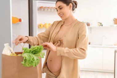 Mutfaktaki açık buzdolabının yanında alışveriş torbası taşıyan hamile bir kadın.