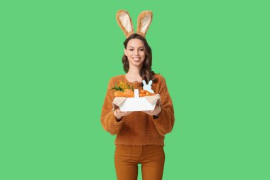 Paskalya tavşanı kulaklıklı mutlu genç bir kadın yeşil arka planda havuç dolu bir sepet tutuyor.