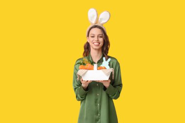 Paskalya tavşanı kulaklıklı mutlu genç kadın sarı arka planda havuçlu sepet tutuyor.