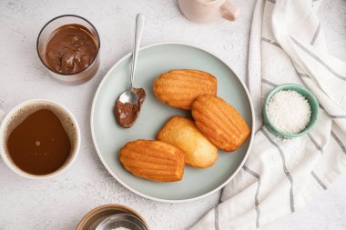 Lezzetli madeleine tabağı, kaşık, bir fincan kahve, bir bardak çikolata kreması ve hafif arka planda hindistan cevizi gevreği.