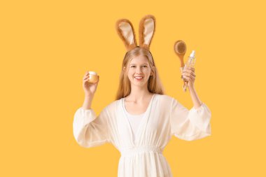 Tavşan kulaklı güzel bir kadın elinde kozmetik ürünler ve sarı arka planda masaj fırçası tutuyor.