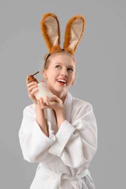 Bornozlu, tavşan kulaklı ve gri arka planda bir şişe kozmetik ürünü olan güzel bir kadın.