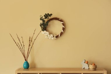 Bunny figurine, eggs and pussy willow branches on dresser near light wall with Easter wreath