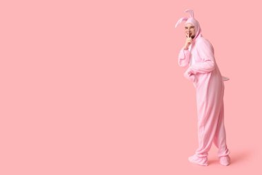 Tavşan kostümlü genç adam pembe arka planda sessizlik jesti gösteriyor. Paskalya kutlaması