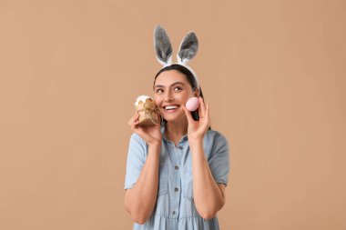 Tavşan kulaklı güzel bir genç kadın. Tatlı paskalya pastası ve kahverengi arka planda yumurta.