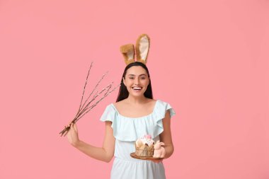Tatlı Paskalya pastası, oyuncak tavşan ve pembe arka planda söğüt dalları olan tavşan kulaklı güzel genç bir kadın.