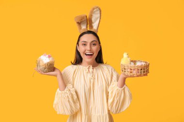 Tavşan kulaklı güzel bir genç kadın. Tatlı paskalya pastası, yumurta ve sarı arka planda bir piliç.