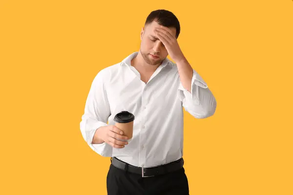 Tired young man with paper cup of coffee on yellow background