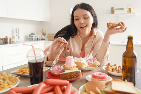 Молодая женщина за столом с нездоровой едой на кухне. Концепция переедания