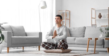 Pijamalı genç adam evde meditasyon yapıyor.