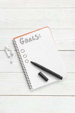 Liste yapmak için boş bir defter, beyaz ahşap arka planda işaretleyici ve kulaklık. Yeni yıl hedefleri