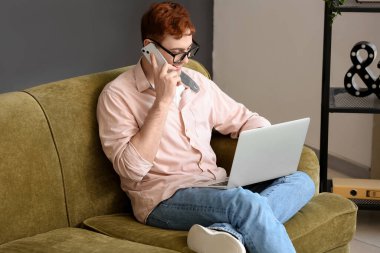 Cep telefonu ve modern dizüstü bilgisayarı olan yakışıklı genç adam internette alışveriş yaparken koltukta oturuyor.