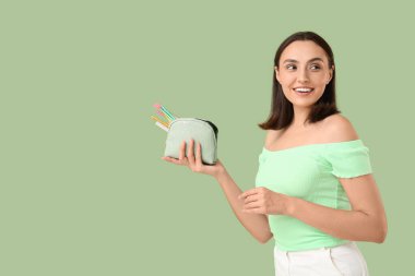 Yeşil arka planda makyaj ürünleriyle dolu kozmetik çantası olan güzel genç bir kadın.