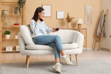 Evdeki kanepede dizüstü bilgisayarı ve bir bardak şarabı olan güzel bir kadın oturuyor.