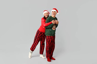 Noel pijamaları ve gri arka planda Noel Baba şapkaları olan mutlu genç bir çiftin portresi.