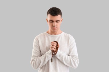 Tespihi olan genç adam gri arka planda dua ediyor.