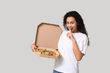 Güzel, genç, mutlu, Afro-Amerikan bir kadın lezzetli pizzasıyla gri arka planda sessiz bir jest yapıyor.