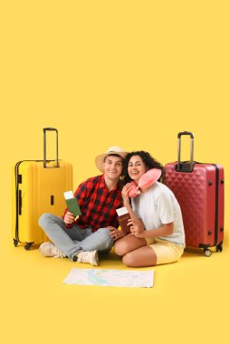 Çantalı, pasaportlu ve sarı arka planda oturan iki turist.