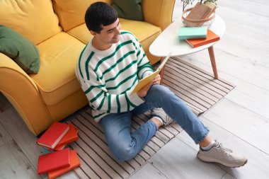 Genç adam evde yerde kitap okuyor.