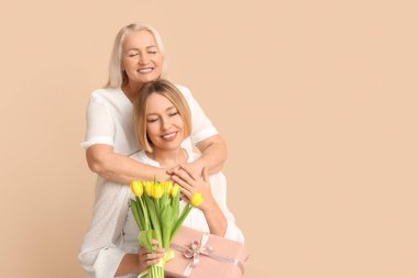 Annesiyle birlikte gülümseyen yetişkin bir kadın, hediye kutusu ve bej arka planda bir buket lale. Uluslararası Kadınlar Günü kutlamaları