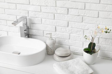 Banyodaki beyaz tuğla duvarın yanındaki tezgahta banyo malzemeleri ve güzel çiçeklerle modern lavabo.
