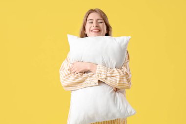 Sarı arka planda pijamalı genç bir kadın yastığa sarılıyor.