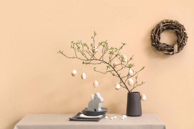 Mutfaktaki bej duvarın yanındaki masada ağaç dalları, paskalya yumurtaları ve tabaklar olan vazo.