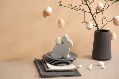 Ağaç dalları olan vazo, bej duvarın yanındaki masada paskalya yumurtası ve tabaklar, yakın plan.