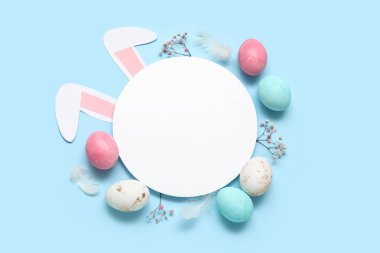 Boş kartla boyanmış Paskalya yumurtaları ve mavi arka planda kağıt tavşan kulakları.