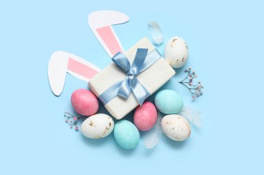 Boyanmış Paskalya yumurtaları, hediye kutusu ve mavi arka planda kağıt tavşan kulakları.