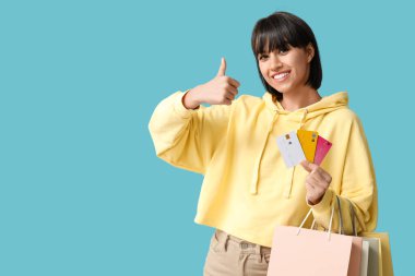 Kredi kartları ve alışveriş çantaları olan güzel genç bir kadın mavi arka planda baş parmak hareketi gösteriyor.