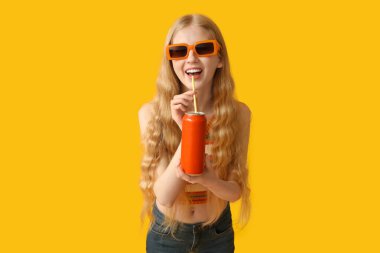 Sarı arka planda bir kutu gazozu olan güneş gözlüklü mutlu genç kadın.