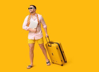 Sarı arka planda dizüstü bilgisayarı ve bavulu olan güneş gözlüklü mutlu bir ofis çalışanı.