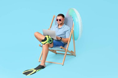 Şişme yüzüklü ve dizüstü bilgisayarlı genç bir ofis çalışanı mavi arka planda şezlongda oturuyor.