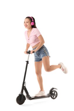 Kulaklıklı güzel bir Asyalı kadın beyaz arka planda modern elektrikli scooter kullanıyor.