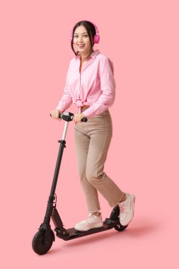 Pembe arka planda modern elektrikli scooter kullanan kulaklıklı mutlu genç Asyalı kadın.