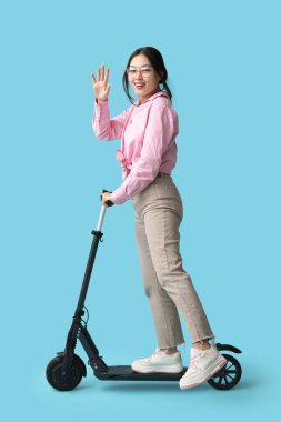 Güzel genç Asyalı kadın mavi arka planda modern elektrikli scooter kullanıyor.