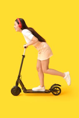Sarı arka planda modern elektrikli scooter kullanan kulaklıklı güzel bir Asyalı kadın.