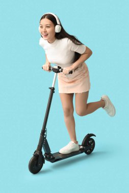 Mavi arka planda modern elektrikli scooter kullanan kulaklıklı mutlu genç Asyalı kadın.