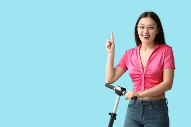Modern elektrikli scooterlı güzel bir Asyalı kadın mavi arka plandaki bir şeyi işaret ediyor.