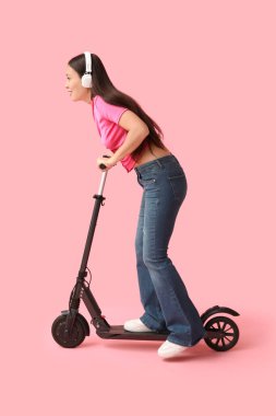 Kulaklıklı güzel bir Asyalı kadın pembe arka planda modern elektrikli scooter kullanıyor.