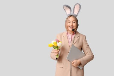 Tavşan kulaklı, kulaklı, dizüstü bilgisayarı ve beyaz arka planda laleleri olan mutlu yetişkin kadın operatör. Paskalya kutlaması