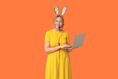 Turuncu arka planda kulaklık ve dizüstü bilgisayarı olan tavşan kulaklı mutlu yetişkin kadın operatör. Paskalya kutlaması
