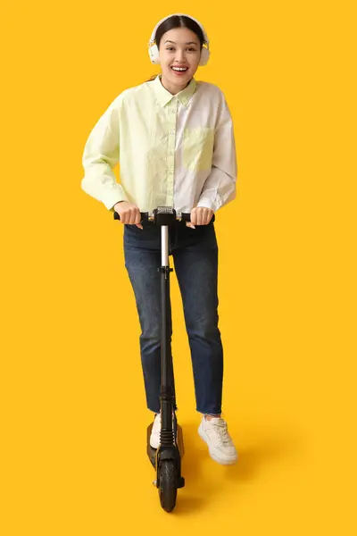 黄色の背景に現代電動キックスクーターに乗ってヘッドフォンで幸せな若いアジアの女性 — ストック写真