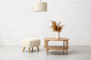 Beyaz tuğla duvarın yanında vazo ve puf kaplı sehpa.