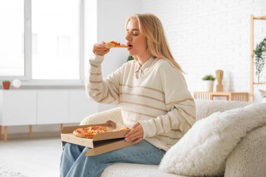 Kanepede oturan ve evde lezzetli pizzalar yiyen genç bir kadın.