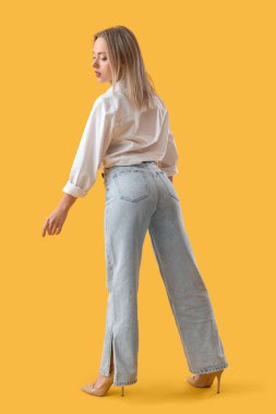 Sarı arka planda kot pantolonlu şık bir genç kadın.