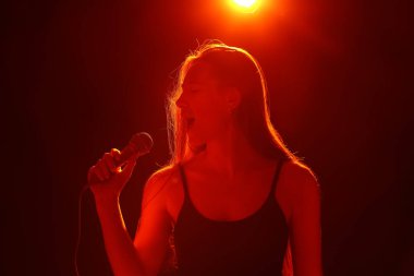 Modern mikrofonlu genç bir kadın karanlık arka planda şarkı söylüyor.