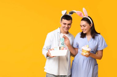 Paskalya tavşanı kulaklı mutlu genç çift, sarı arka planda sepet ve pasta.
