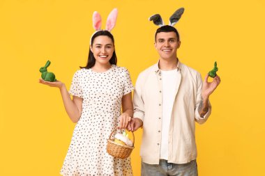 Paskalya tavşanı kulaklı, sepetli ve sarı arka planda oyuncak tavşanları olan mutlu bir çift.